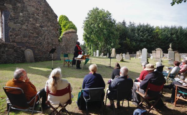 The service held by Newport-on-Tay Church of Scotland for the 900th anniversary of a church being on the location. It is pictured being held outside near the old church ruin on a sunny day. The minister faces the congregation who are sitting in portable chairs.