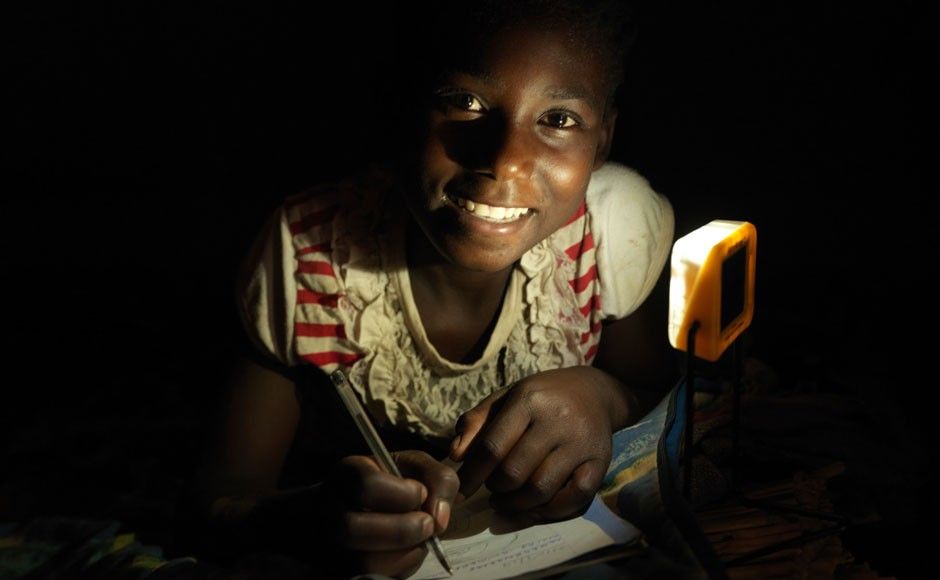 A child writing on paper with the room being partially lit by a light. The child is writing on the paper with a pen and smiling at the camera.