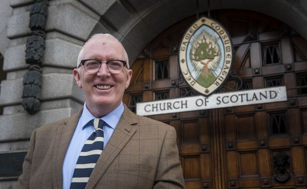 Rev Dr Martin Fair standing in front of the Church of Scotland emblem